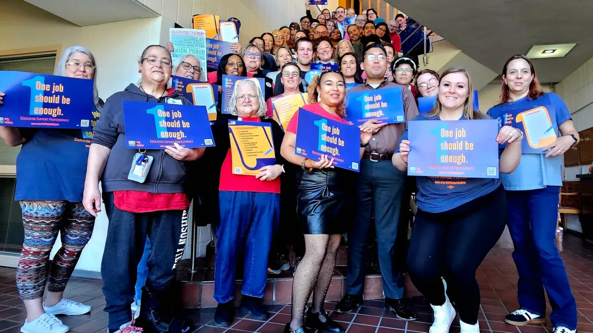 A group of ESPs pose together in the KNEA building in Topeka holding blue signs that say, "One job should be enough."