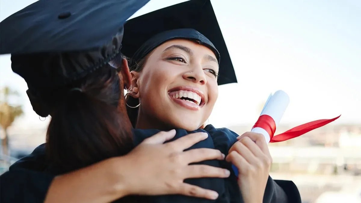 Woman in cap and gown holding diploma and receiving a hug.